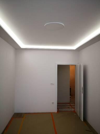 Pusty pokój z białymi ścianami, oświetleniem LED w suficie i okrągłą lampą sufitową, podłoga zabezpieczona folią malarską, otwarte drzwi do kolejnego pomieszczenia.