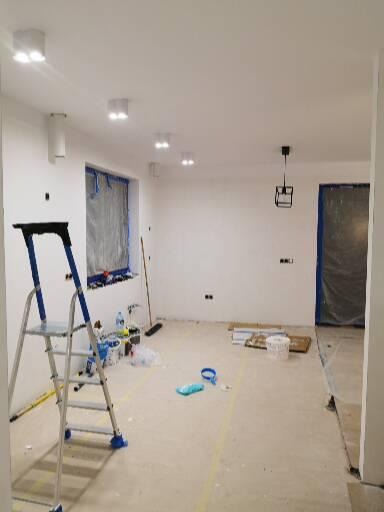 Wnętrze w trakcie prac wykończeniowych: zabezpieczone okno folią, rozstawiona drabina, widoczne narzędzia i materiały malarskie, surowa podłoga przygotowana do dalszych prac.