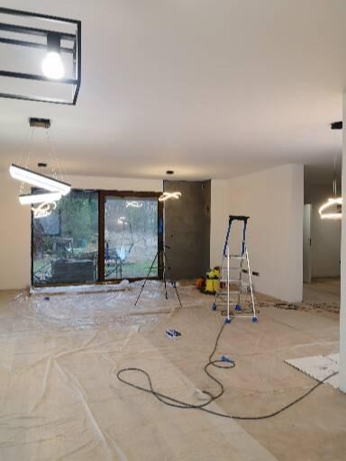 Przygotowania do prac wykończeniowych w przestronnym salonie: podłoga zabezpieczona folią, drabina malarska, statyw oświetleniowy i odkurzacz widoczne w tle. Nowoczesne oświetlenie sufitowe.