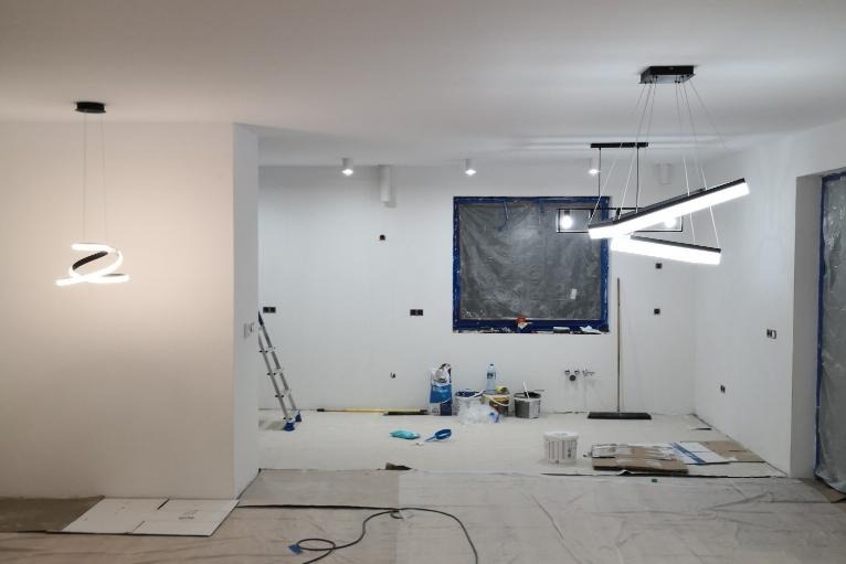 Wnętrze w trakcie prac przygotowawczych: zabezpieczone okno folią, podłoga zakryta, widoczne wiadra z farbą i drabina, nowoczesne lampy sufitowe.