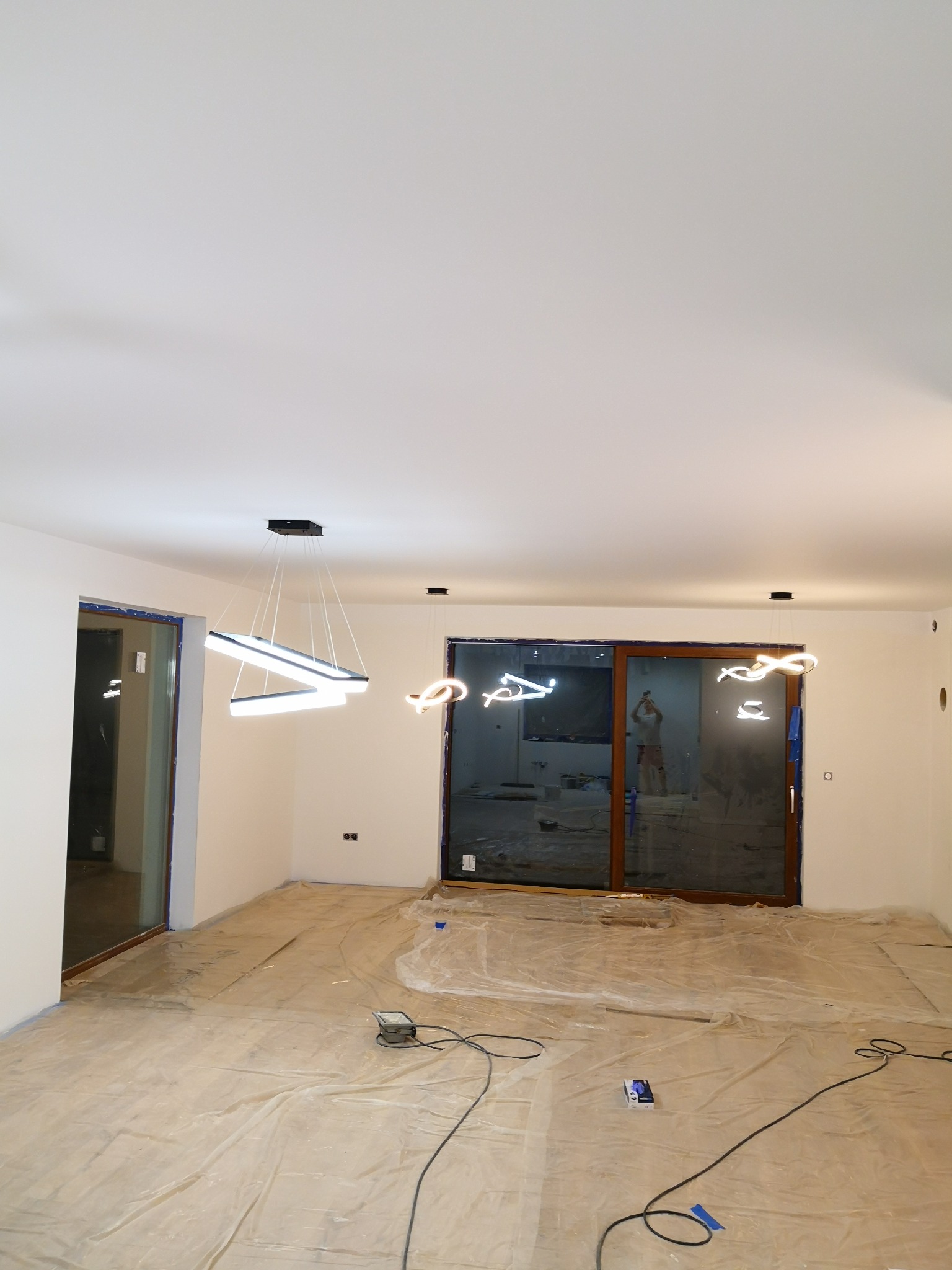 Wnętrze pokoju w trakcie remontu, podłoga zabezpieczona folią, zamontowane nowoczesne lampy LED, widoczne okna przesuwne.