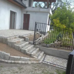 Granitowe schody prowadzące do domu, z kostki ułożonej w łukowaty wzór, obok ozdobne metalowe ogrodzenie z motywem liści winogron.