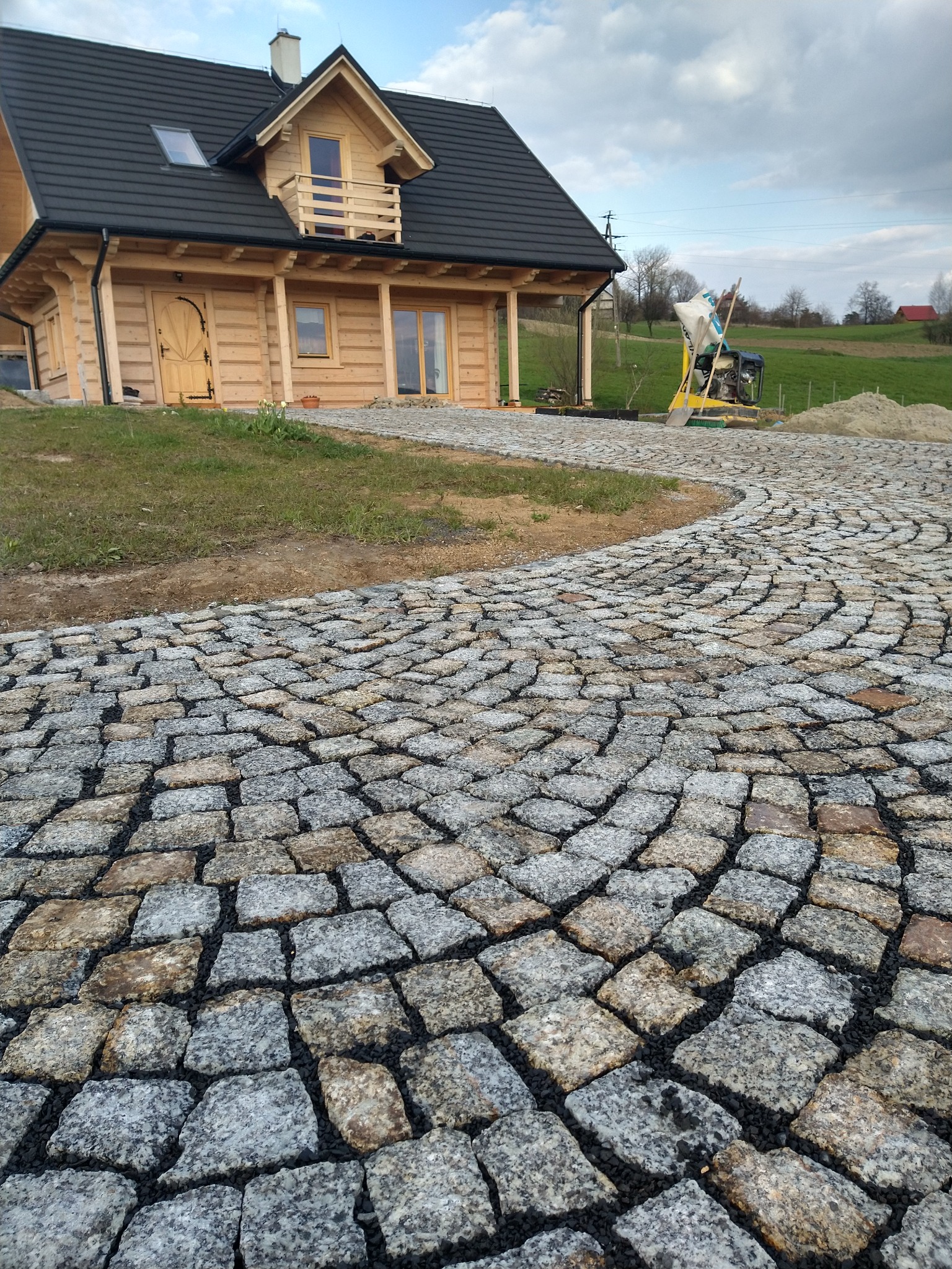 Zakrzywiona ścieżka z kostki granitowej prowadząca do drewnianego domu z balkonem, z widoczną ubijarką w tle.