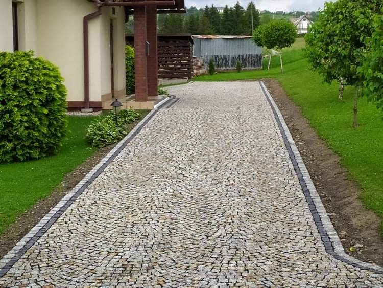 Ułożona kostka granitowa w odcieniach szarości i beżu tworząca wzór wachlarza na podjeździe, obrzeża z ciemniejszej kostki, trawnik i budynek w tle.
