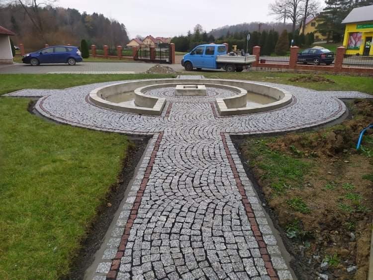 Ogród z fontanną otoczoną ścieżką z szarej kostki, obrzeża z czerwonej kostki, w tle budynki i samochody.