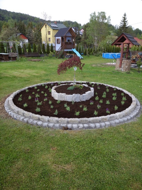 Dwupoziomowy klomb z kostki granitowej w okrągłym kształcie, obsadzony młodymi roślinami, na tle zielonego trawnika, placu zabaw i zabudowań.