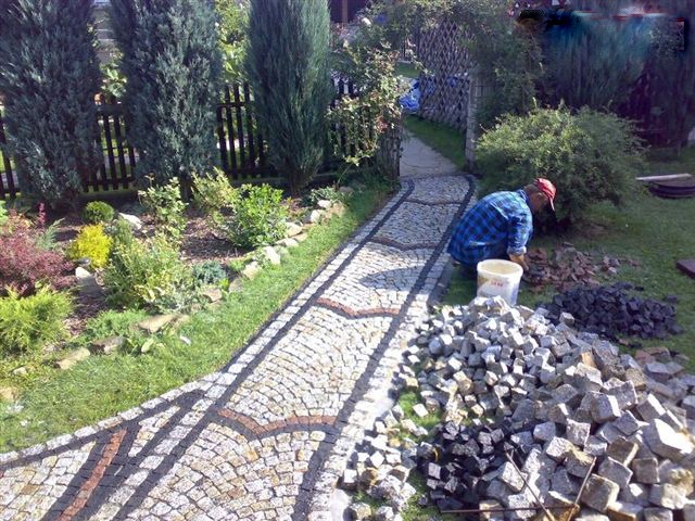 Pracownik w czerwonej czapce układa wzorzystą ścieżkę z kostki brukowej w ogrodzie, widoczne kopce kostki w dwóch kolorach i wiadro z zaprawą.