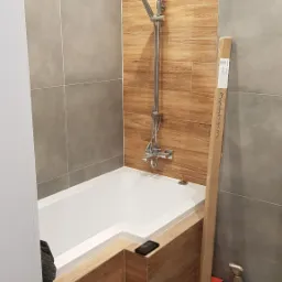 Nowoczesna łazienka z wanną obudowaną drewnopodobnymi płytkami, chromowaną baterią prysznicową z deszczownicą i słuchawką, szare płytki na ścianach i podłodze, kartonowe opakowanie opierające się...