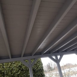 GregBud - Szara drewniana pergola widziana od dołu, z widocznym zadaszeniem z desek i konstrukcją wsporczą, w tle zimowy krajobraz z zielonymi krzewami.