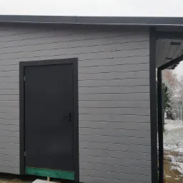 Szara, drewniana budowla z ciemnymi elementami wykończeniowymi, takimi jak drzwi i rynny, na tle zimowego krajobrazu z widocznym śniegiem.