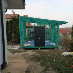 GregBud - Szkielet nowej, pomalowanej na zielono konstrukcji z czarnymi drzwiami, prawdopodobnie szopy lub małego budynku gospodarczego, w trakcie budowy obok domu jednorodzinnego.