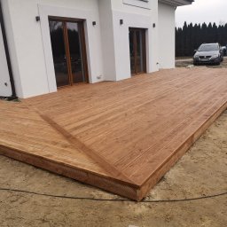 GregBud - Nowoczesny taras drewniany przylegający do białego domu z dużymi oknami, widok na auto i kontener na śmieci w tle.