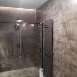 GregBud - Szklana kabina prysznicowa z czarną ramą w łazience z płytkami imitującymi beton, widoczne prace wykończeniowe, w tle toaleta i narzędzia.