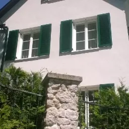 Elewacja dwupiętrowego domu z białego tynku z zielonymi okiennicami, widoczna zza metalowego ogrodzenia i żywopłotu.