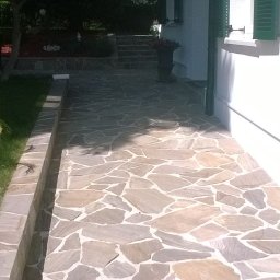 GregBud - Kamienna ścieżka z nieregularnych płyt prowadząca wzdłuż białego budynku z zielonymi okiennicami, obok trawnika z palmą w białej donicy.