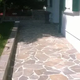 Kamienna ścieżka z nieregularnych płyt prowadząca wzdłuż białego budynku z zielonymi okiennicami, obok trawnika z palmą w białej donicy.