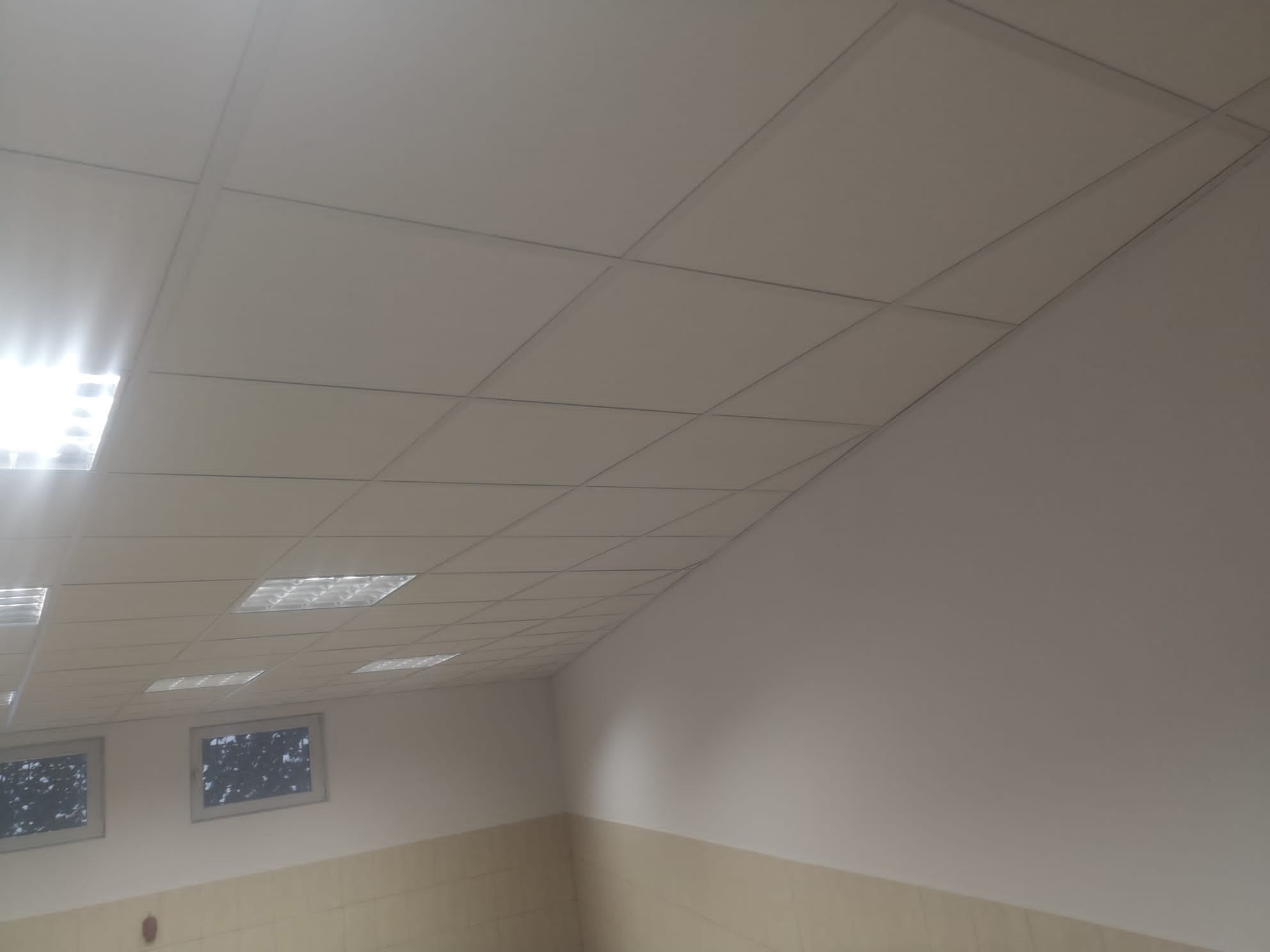 Sufit podwieszany z kasetonów i oświetleniem rastrowym w biurze, widok z dołu pod kątem, ściany pomalowane na biało z dolnym pasem w kolorze beżowym.