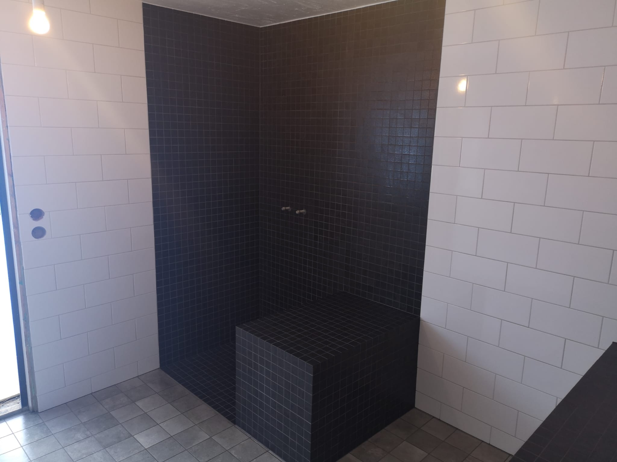 Nowoczesna kabina prysznicowa z czarnymi mozaikowymi płytkami i wbudowanym siedziskiem, kontrastująca z białymi, prostokątnymi płytkami na ścianach bocznych. Szare płytki podłogowe i surowy...