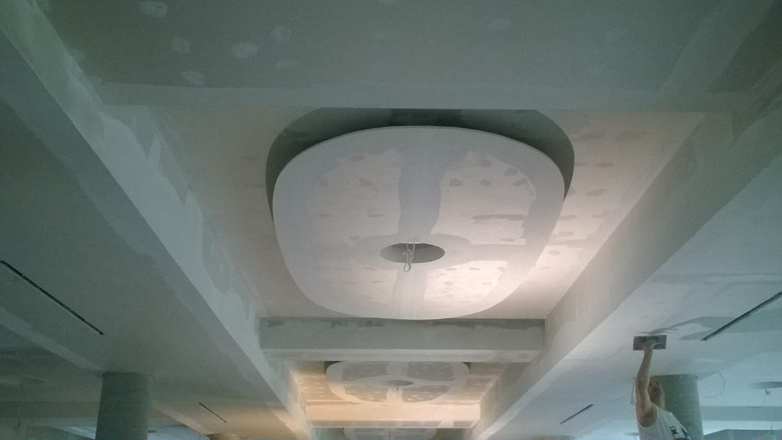 Sufit podwieszany z owalnymi elementami dekoracyjnymi w trakcie montażu, widoczny pracownik szpachlujący połączenia płyt gipsowo-kartonowych.