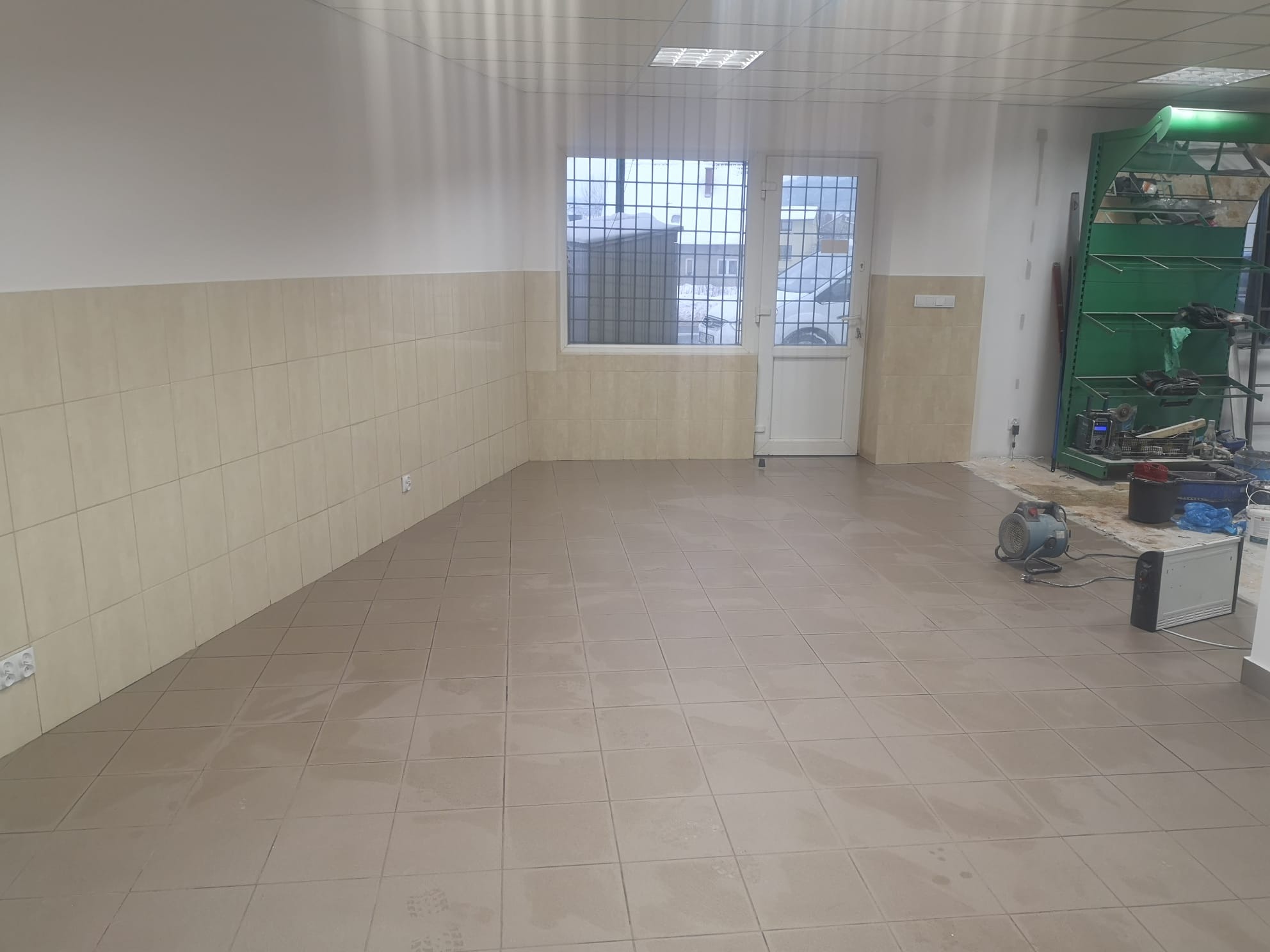 Wnętrze komercyjne z beżowymi płytkami podłogowymi i ściennymi, widoczne narzędzia budowlane, grzejnik i metalowy regał, prawdopodobnie w trakcie prac wykończeniowych.