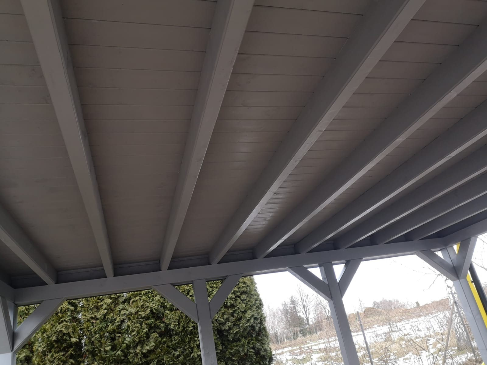 Szara drewniana pergola widziana od dołu, z widocznym zadaszeniem z desek i konstrukcją wsporczą, w tle zimowy krajobraz z zielonymi krzewami.