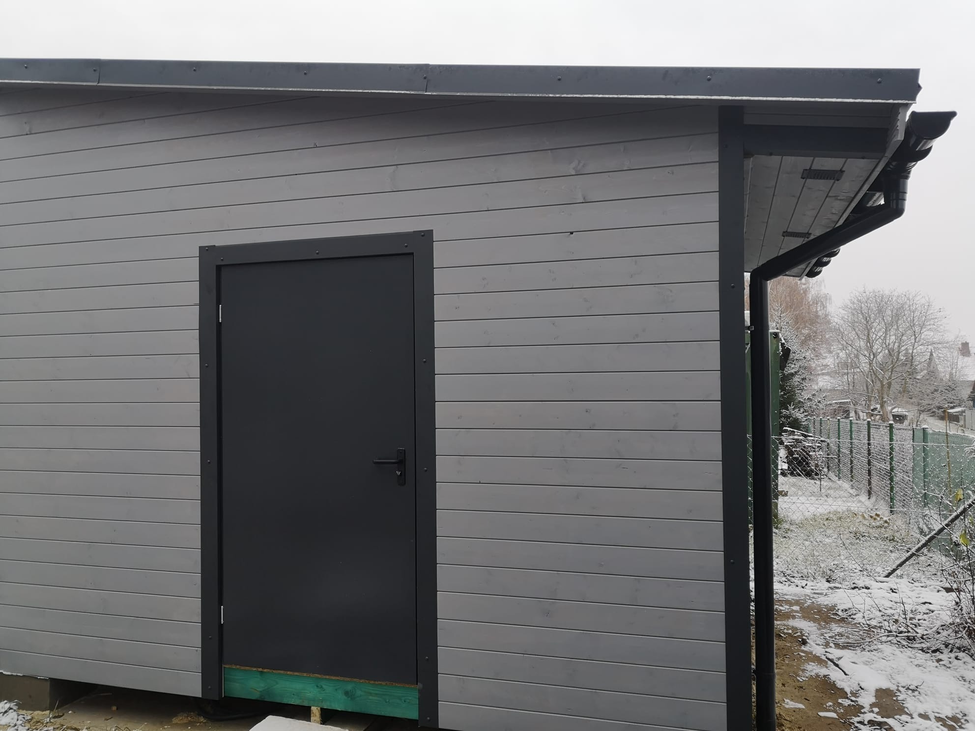 Szara, drewniana budowla z ciemnymi elementami wykończeniowymi, takimi jak drzwi i rynny, na tle zimowego krajobrazu z widocznym śniegiem.