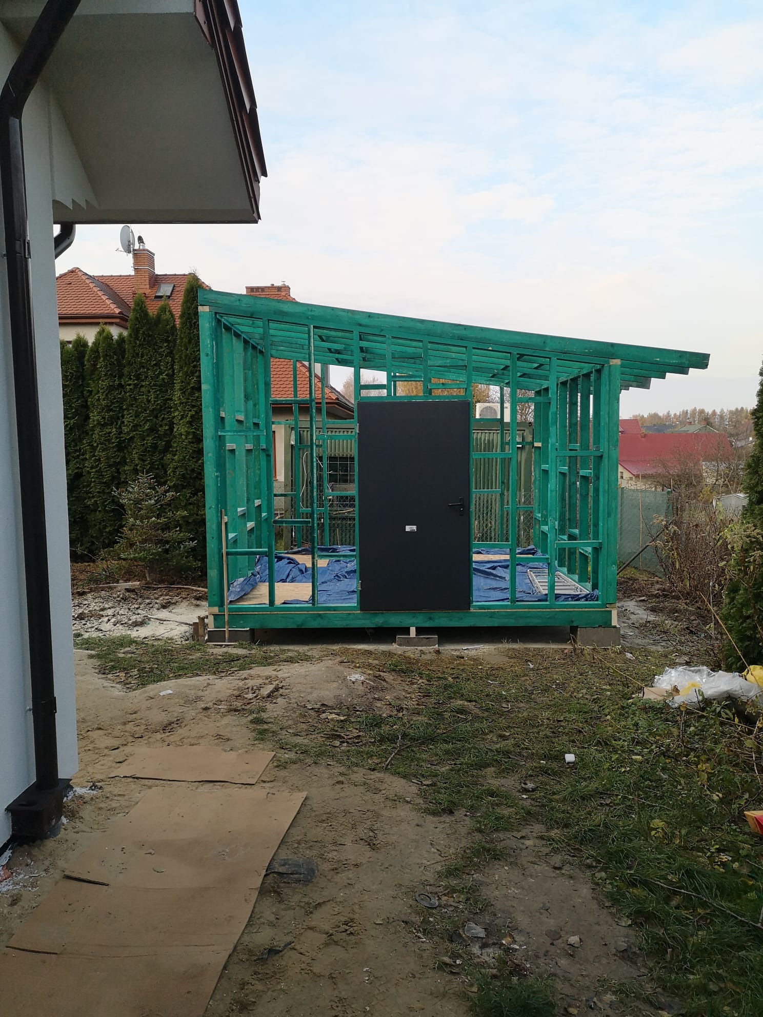 Szkielet nowej, pomalowanej na zielono konstrukcji z czarnymi drzwiami, prawdopodobnie szopy lub małego budynku gospodarczego, w trakcie budowy obok domu jednorodzinnego.