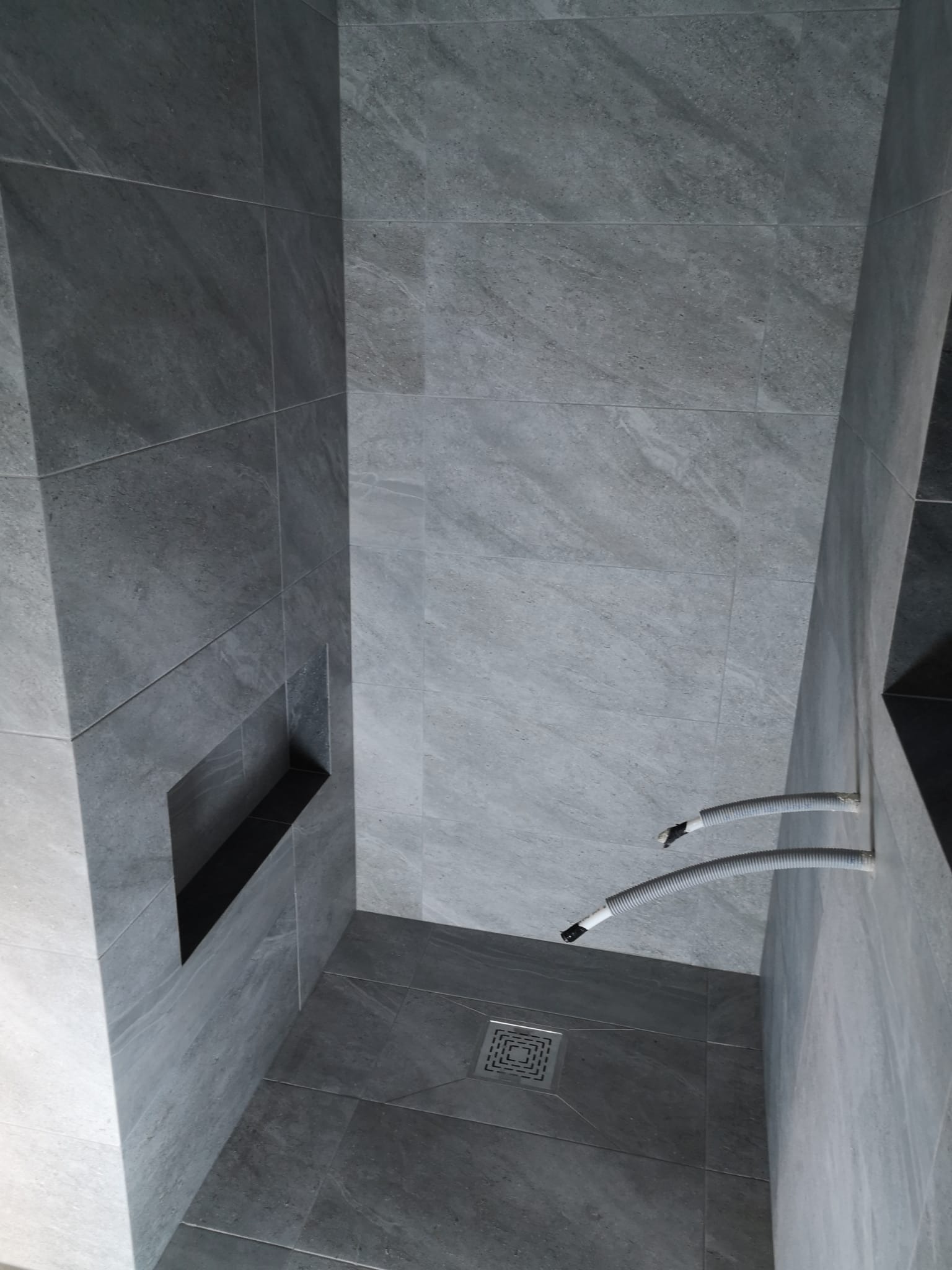 Szara łazienka z płytkami imitującymi kamień, widoczna wnęka w ścianie i rury instalacyjne przed zabudową, odpływ liniowy w podłodze.