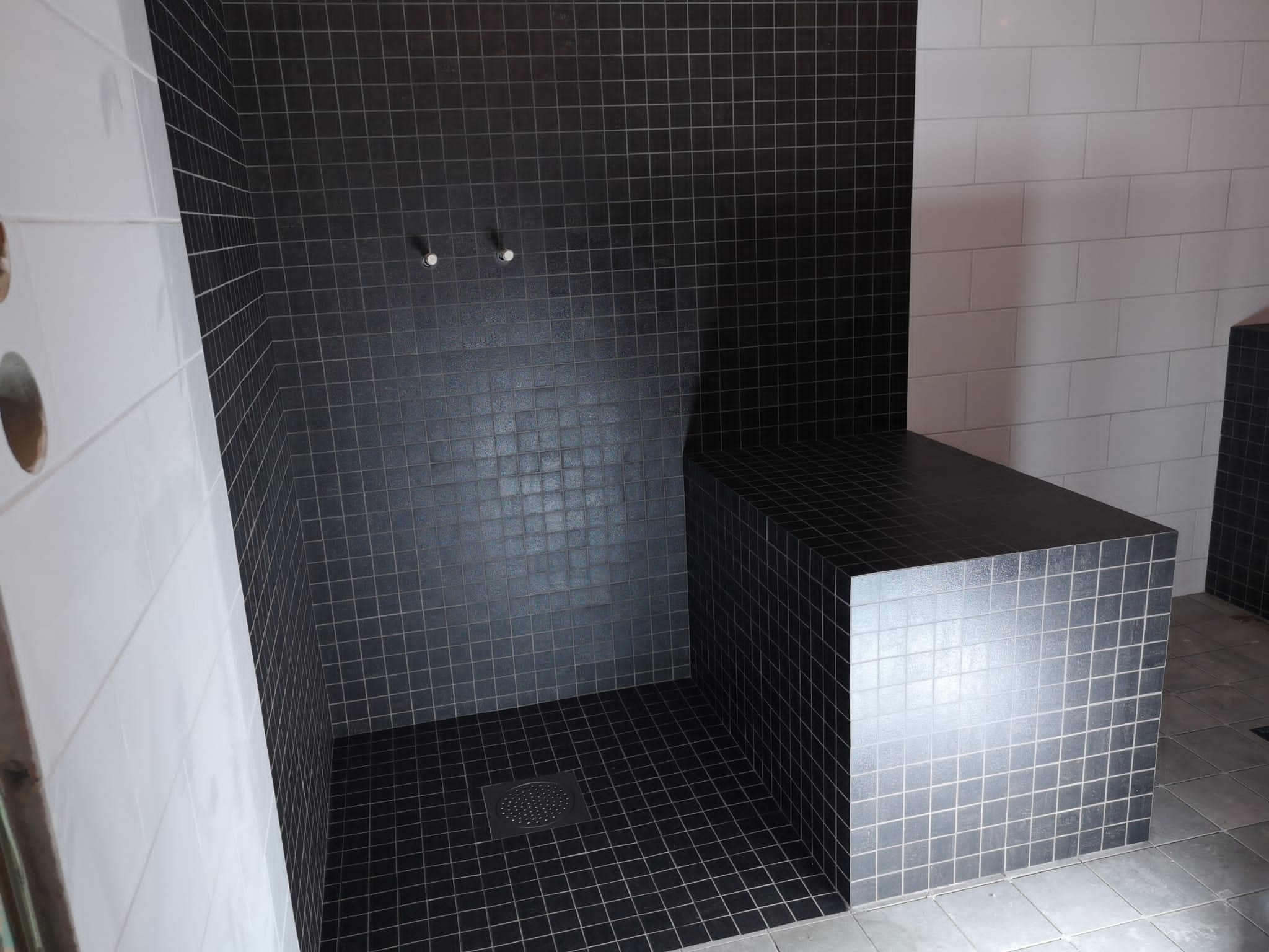 Czarna kabina prysznicowa wyłożona mozaiką z siedziskiem, widoczne dwa uchwyty na słuchawkę prysznicową i odpływ liniowy, kontrastująca z białą ścianą wyłożoną dużymi płytkami.