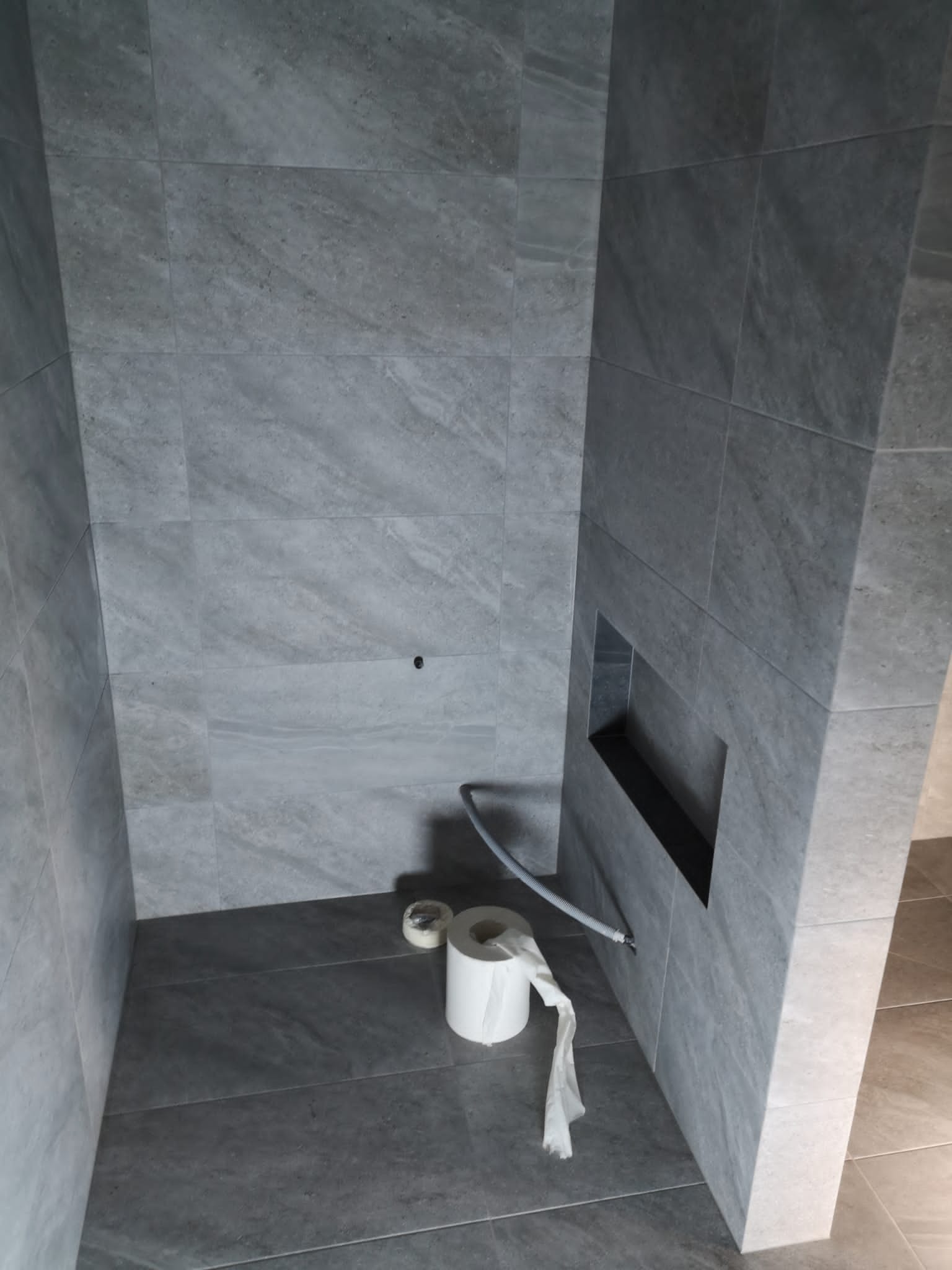 Szara, wykafelkowana kabina prysznicowa z wnęką w ścianie, z rolką papieru toaletowego na podłodze i wystającym wężem.