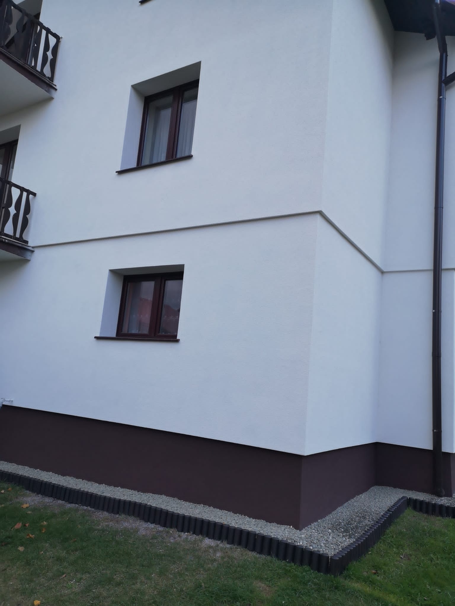 Fragment elewacji budynku z białym tynkiem i brązowym cokołem, widoczne dwa okna z brązowymi ramami, rynna oraz obrzeże z palisady i jasnego żwiru.