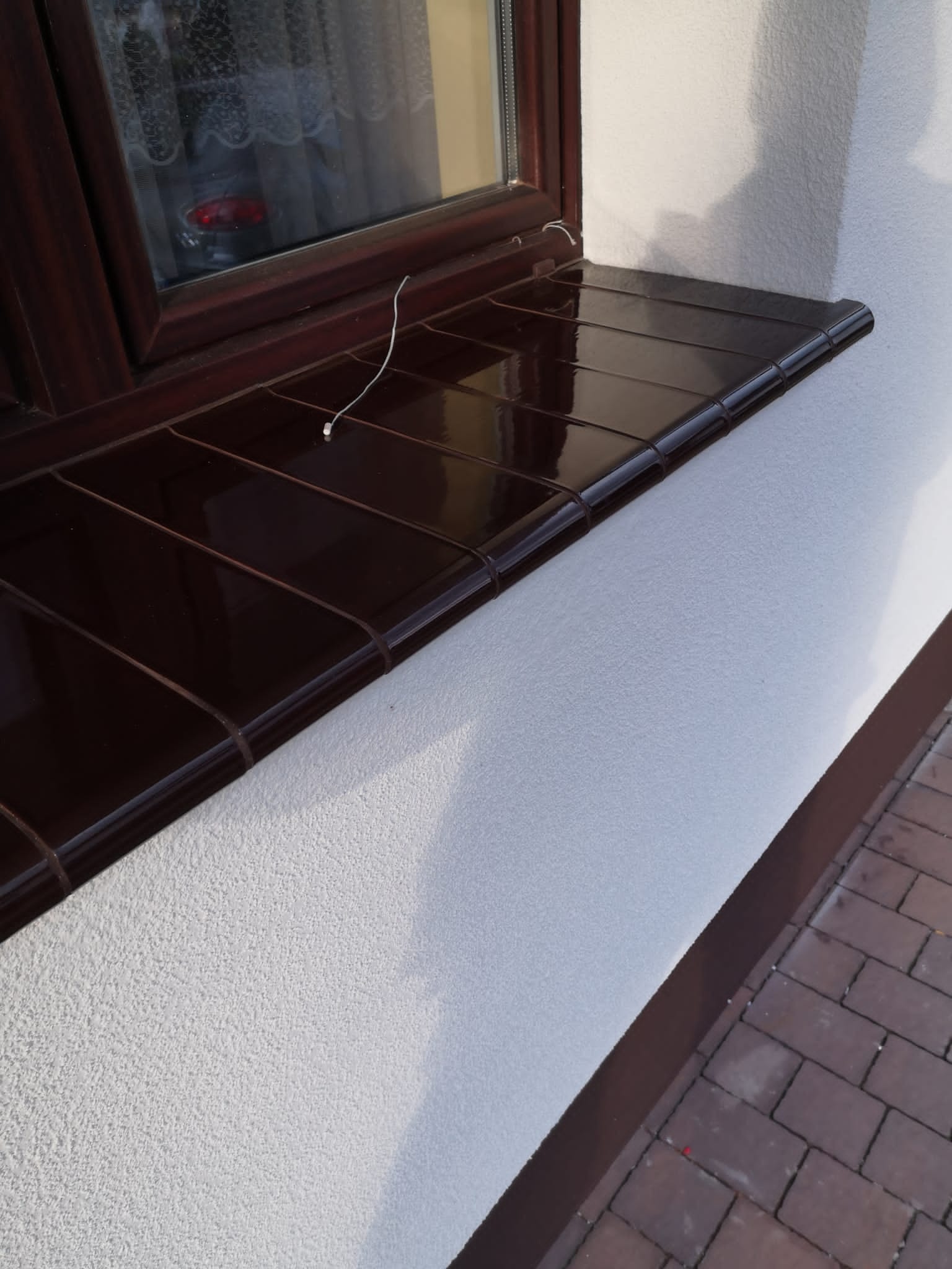 Brązowy, ceramiczny parapet zewnętrzny pod oknem, z widoczną fakturą tynku na elewacji budynku.
