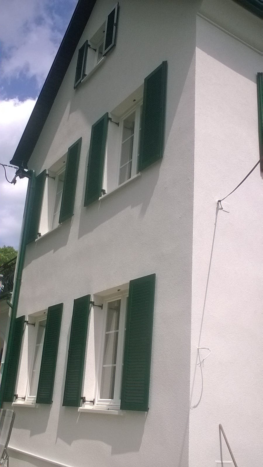 Boczny widok odnowionego budynku z białymi ścianami i zielonymi okiennicami, widoczne przewody na elewacji.