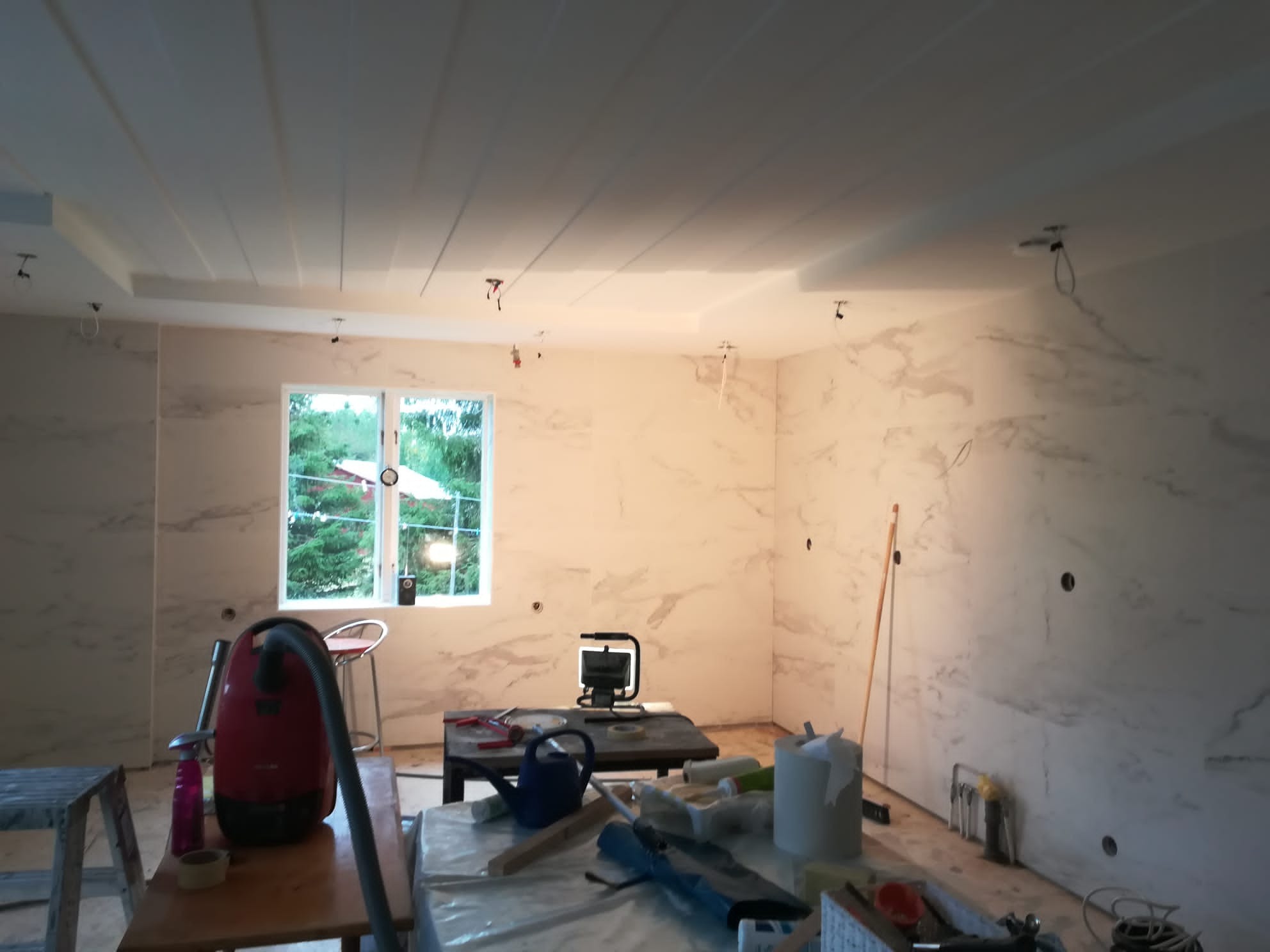 Pokój w trakcie prac wykończeniowych: ściany wyłożone panelami imitującymi marmur, odkryte przewody oświetleniowe zwisające z sufitu, narzędzia i akcesoria malarskie na stole, widok na zieleń przez...