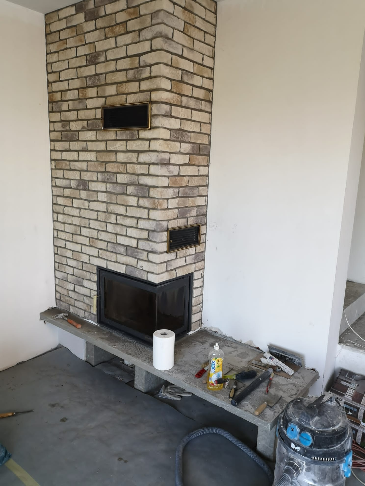 Wnętrze w trakcie wykańczania z obudowanym kominkiem z cegły klinkierowej, widoczne narzędzia i akcesoria budowlane, odkurzacz przemysłowy oraz rolka papieru.