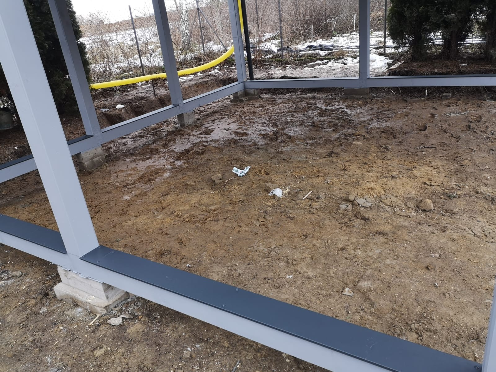 Stalowa konstrukcja szkieletowa w trakcie budowy, widoczny fundament i fragment żółtego przewodu w ziemi.