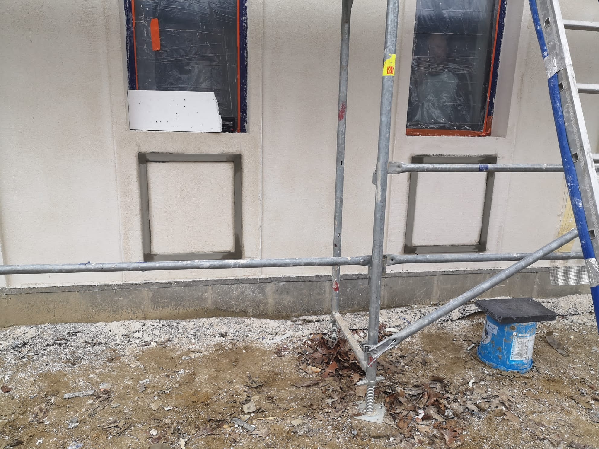 Elewacja budynku w trakcie ocieplania, widoczne rusztowanie, okna zabezpieczone folią, fragment ocieplenia i grunt pod budynkiem.