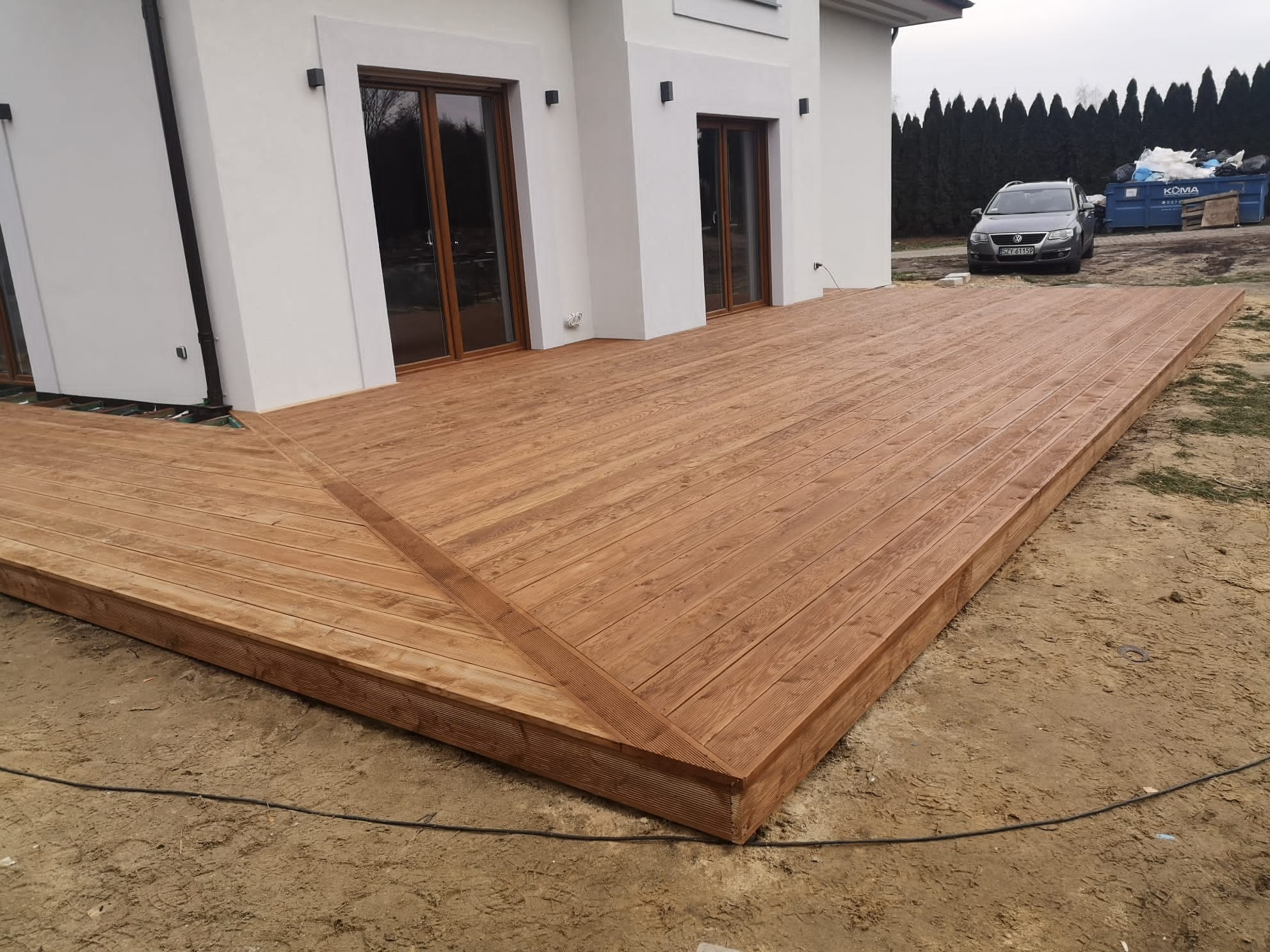 Nowoczesny taras drewniany przylegający do białego domu z dużymi oknami, widok na auto i kontener na śmieci w tle.