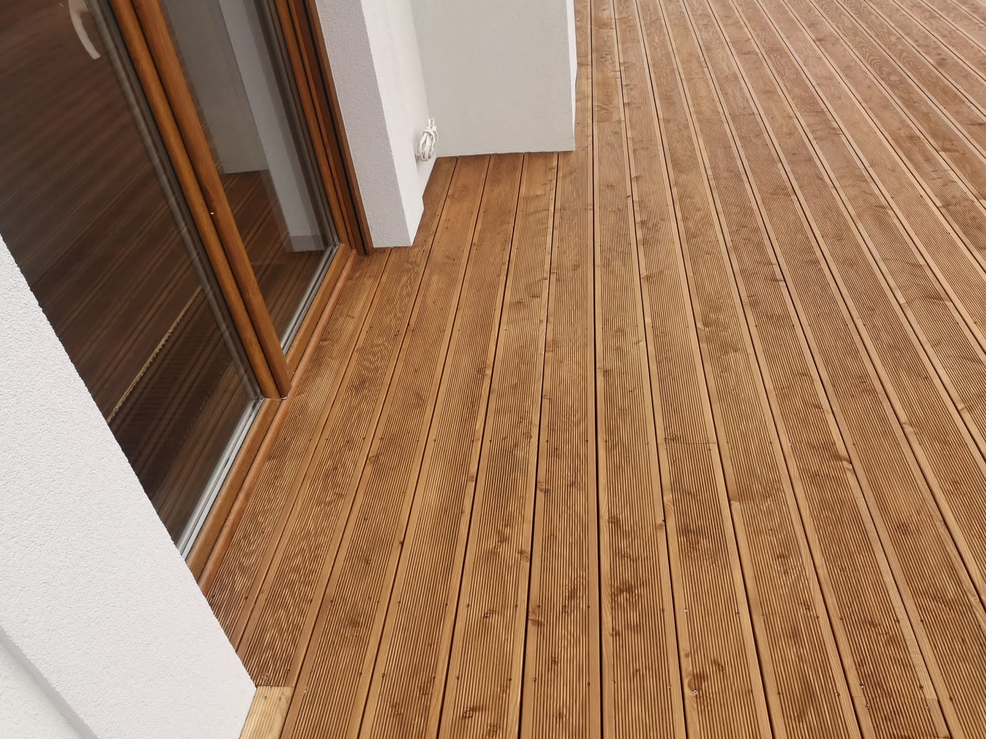 Nowy taras z desek ryflowanych, ułożony przy białej ścianie budynku z oknem balkonowym, widok z góry pod kątem.