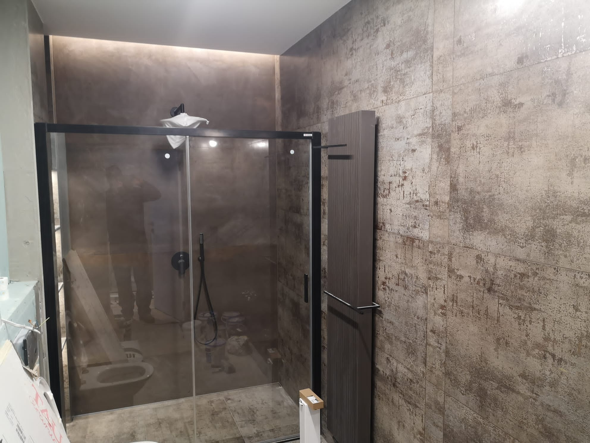 Szklana kabina prysznicowa z czarną ramą w łazience z płytkami imitującymi beton, widoczne prace wykończeniowe, w tle toaleta i narzędzia.