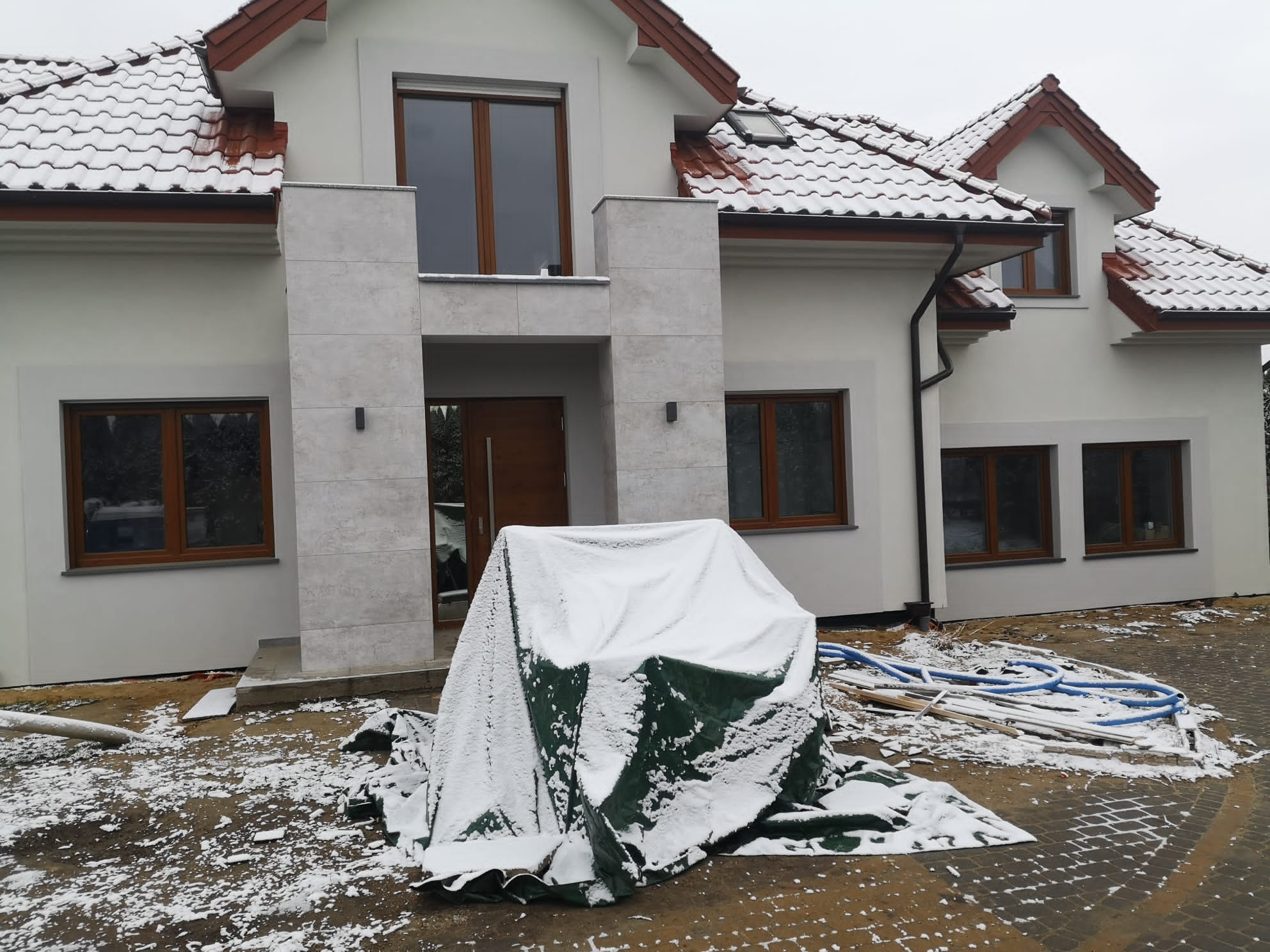 Nowoczesny dom jednorodzinny w Rychwałdzie z elementami wykończenia elewacji, widoczne okna z brązowymi ramami i pokryty śniegiem teren budowy z materiałami budowlanymi.