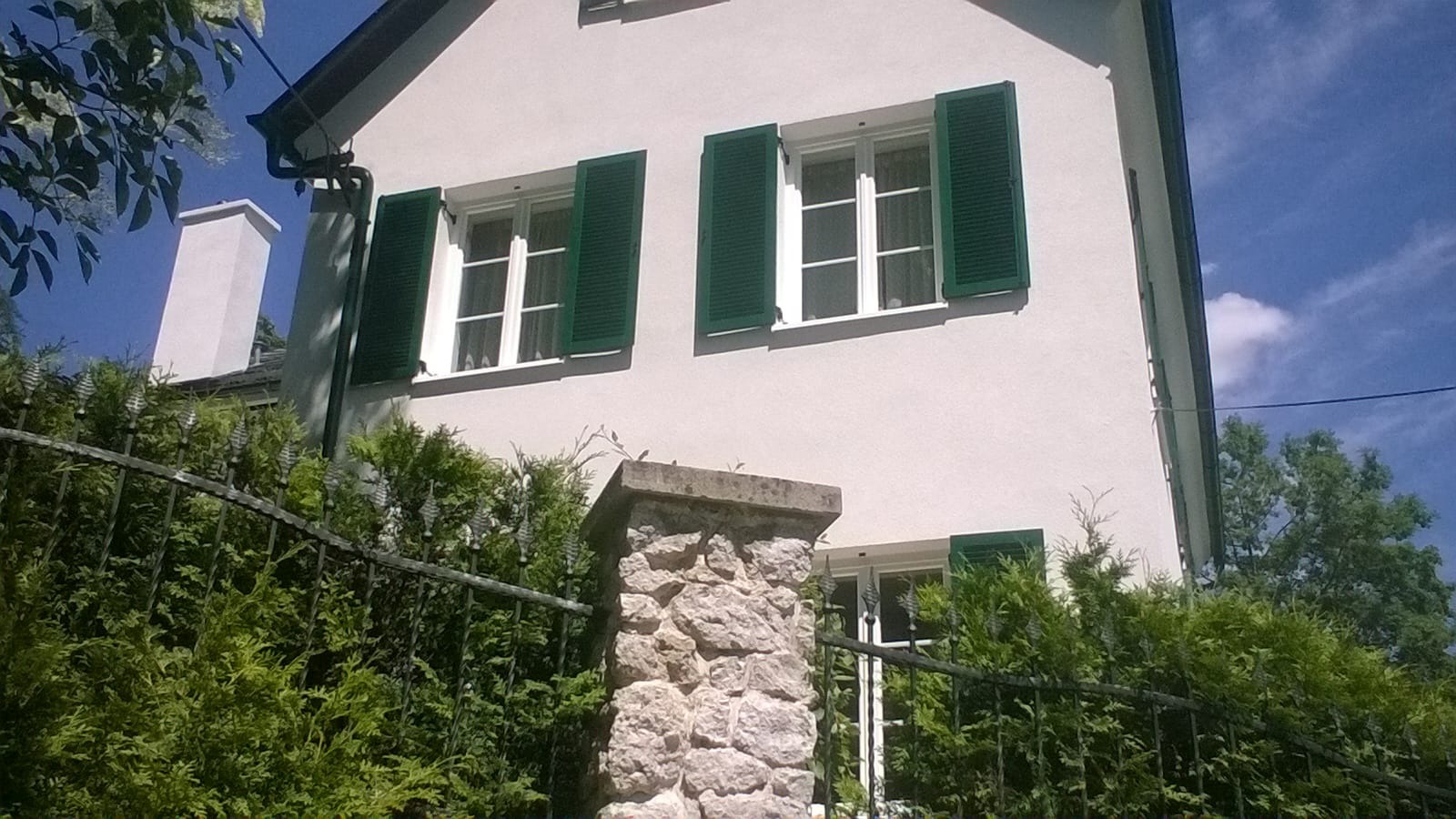 Elewacja dwupiętrowego domu z białego tynku z zielonymi okiennicami, widoczna zza metalowego ogrodzenia i żywopłotu.