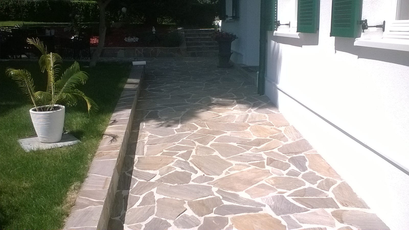 Kamienna ścieżka z nieregularnych płyt prowadząca wzdłuż białego budynku z zielonymi okiennicami, obok trawnika z palmą w białej donicy.