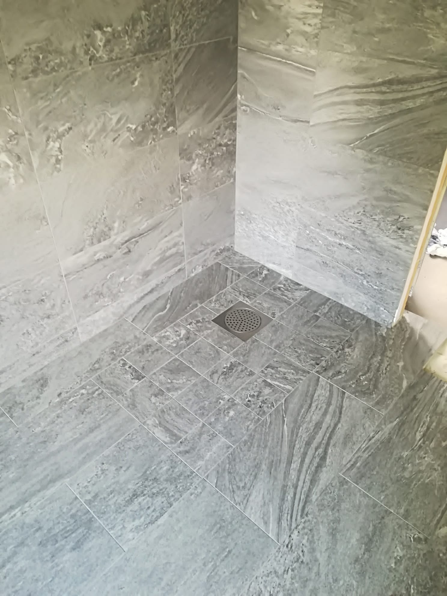 Wykończony brodzik z szarych, marmurkowych płytek ceramicznych, z odpływem liniowym w rogu.