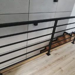 Nowoczesna balustrada schodowa z czarnych, poziomych profili stalowych na drewnianych schodach, z tłem z szarych, kwadratowych płytek ściennych.