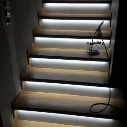 Oświetlone schody drewniane z taśmami LED między stopniami, widoczny fragment instalacji elektrycznej i narzędzie monterskie na jednym ze stopni.