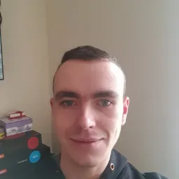 Portret mężczyzny z krótkimi włosami i niebieskimi oczami, ubrany w czarną bluzę z kapturem, na tle jasnej ściany i pudełek.