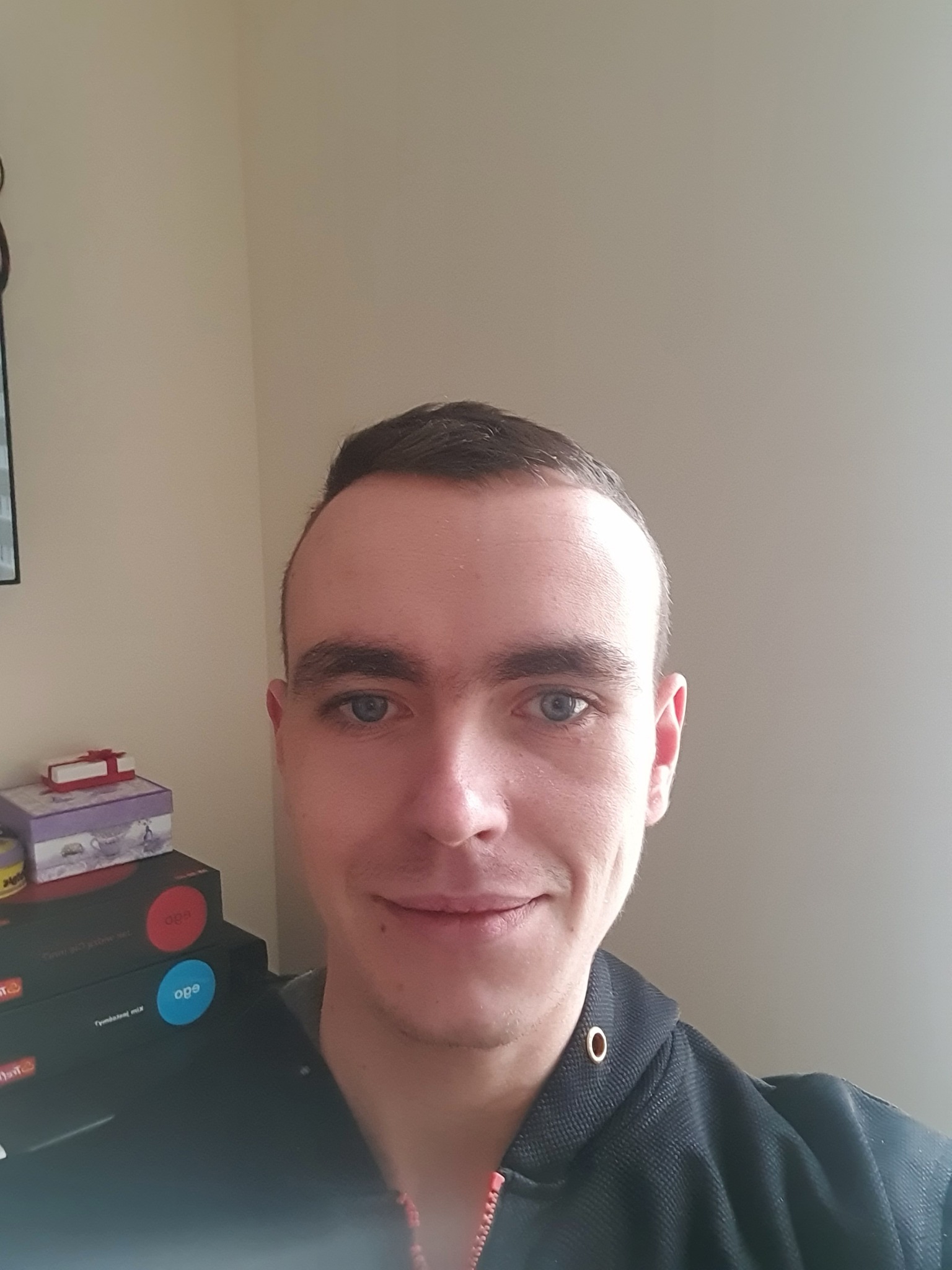 Portret mężczyzny z krótkimi włosami i niebieskimi oczami, ubrany w czarną bluzę z kapturem, na tle jasnej ściany i pudełek.