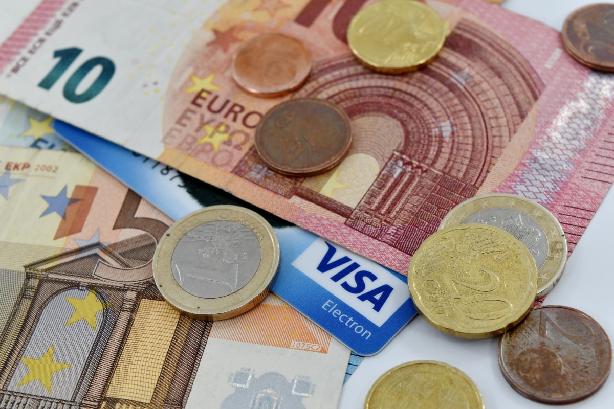 Rozrzucone euro banknoty o nominałach 5, 10 i 50, monety euro o różnych nominałach oraz karta Visa Electron na białym tle.
