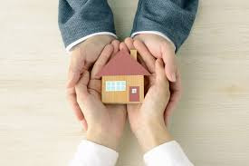 Dłonie kobiety i mężczyzny trzymają model drewnianego domu z czerwonym dachem na jasnym drewnianym tle.